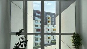 Een kamer met een groot raam dat uitzicht biedt op een meerlaags woongebouw. Op de vloer staan twee potplanten.