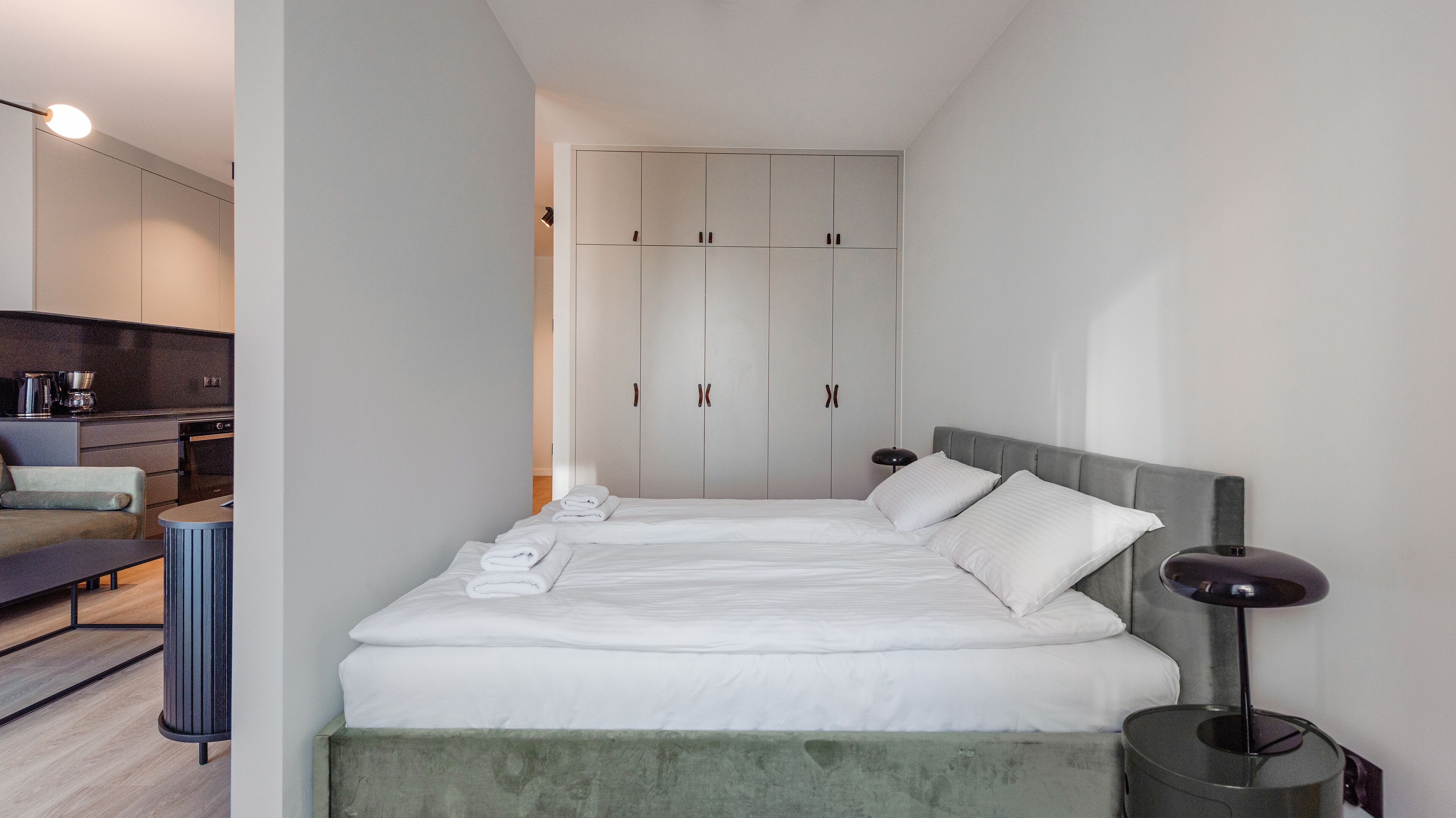 A modern hotel room with a large bed, a bedside table, a lamp, and a view of the kitchen area.