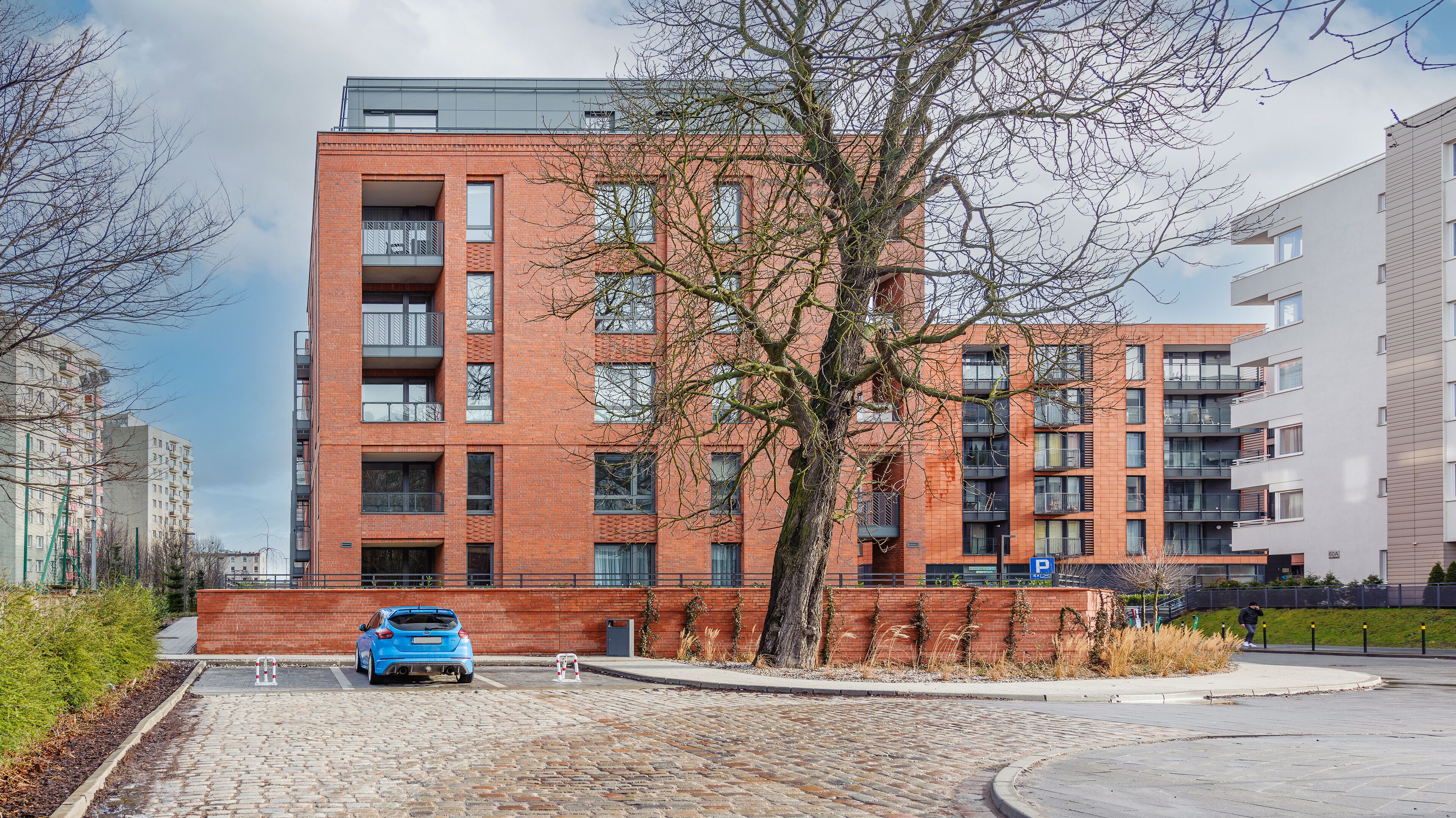 A multi-story residential building with a brick facade, multiple balconies, and a parking area with a blue car.