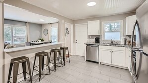 A modern kitchen with white cabinets, a stainless steel refrigerator, and a built-in dishwasher. There are two bar stools and a window with blinds.
