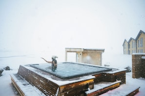 A snow-covered outdoor hot tub area with a wooden bench and a small building in the background.