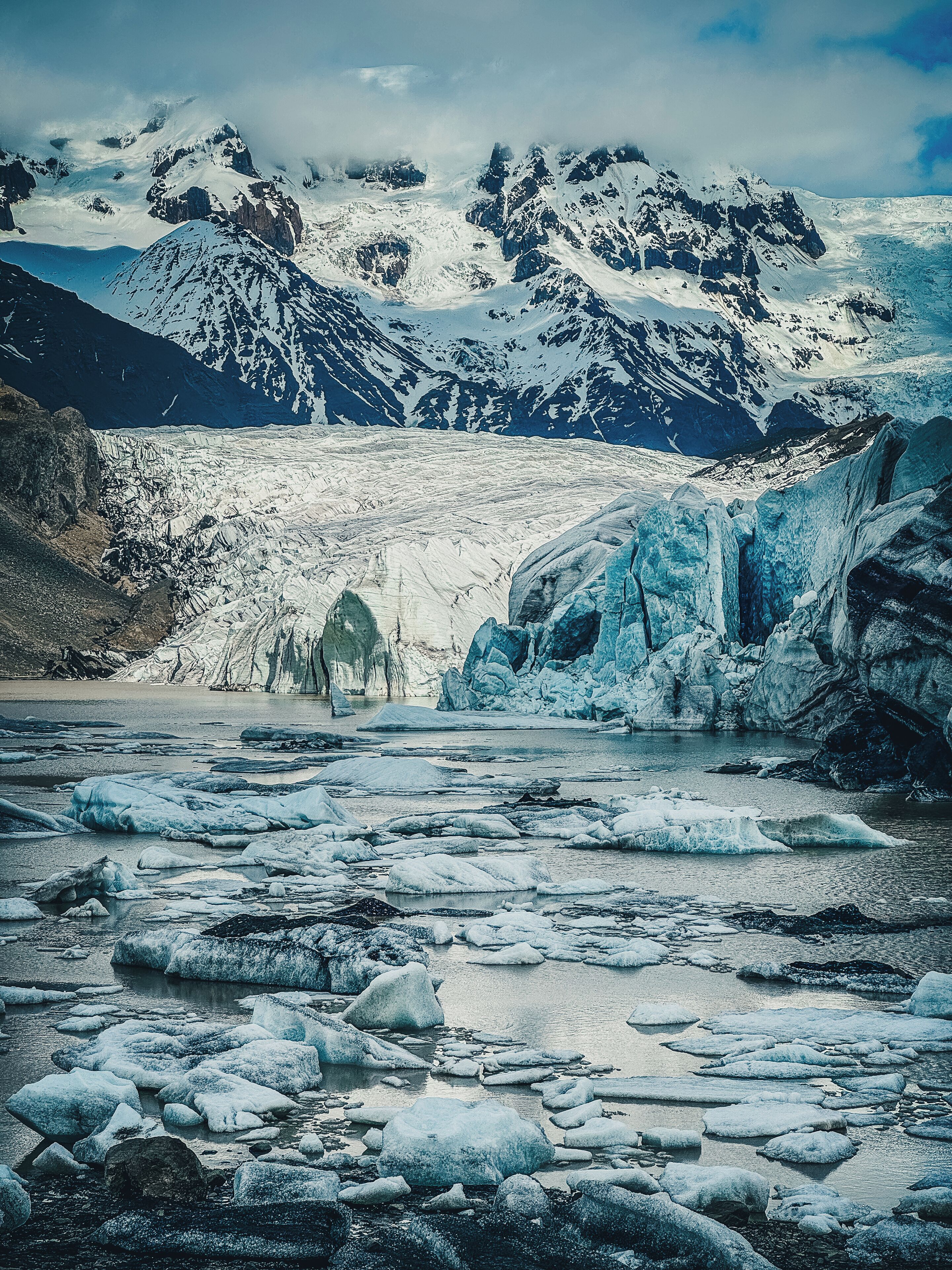 A glacier with icebergs in a river, surrounded by mountains.