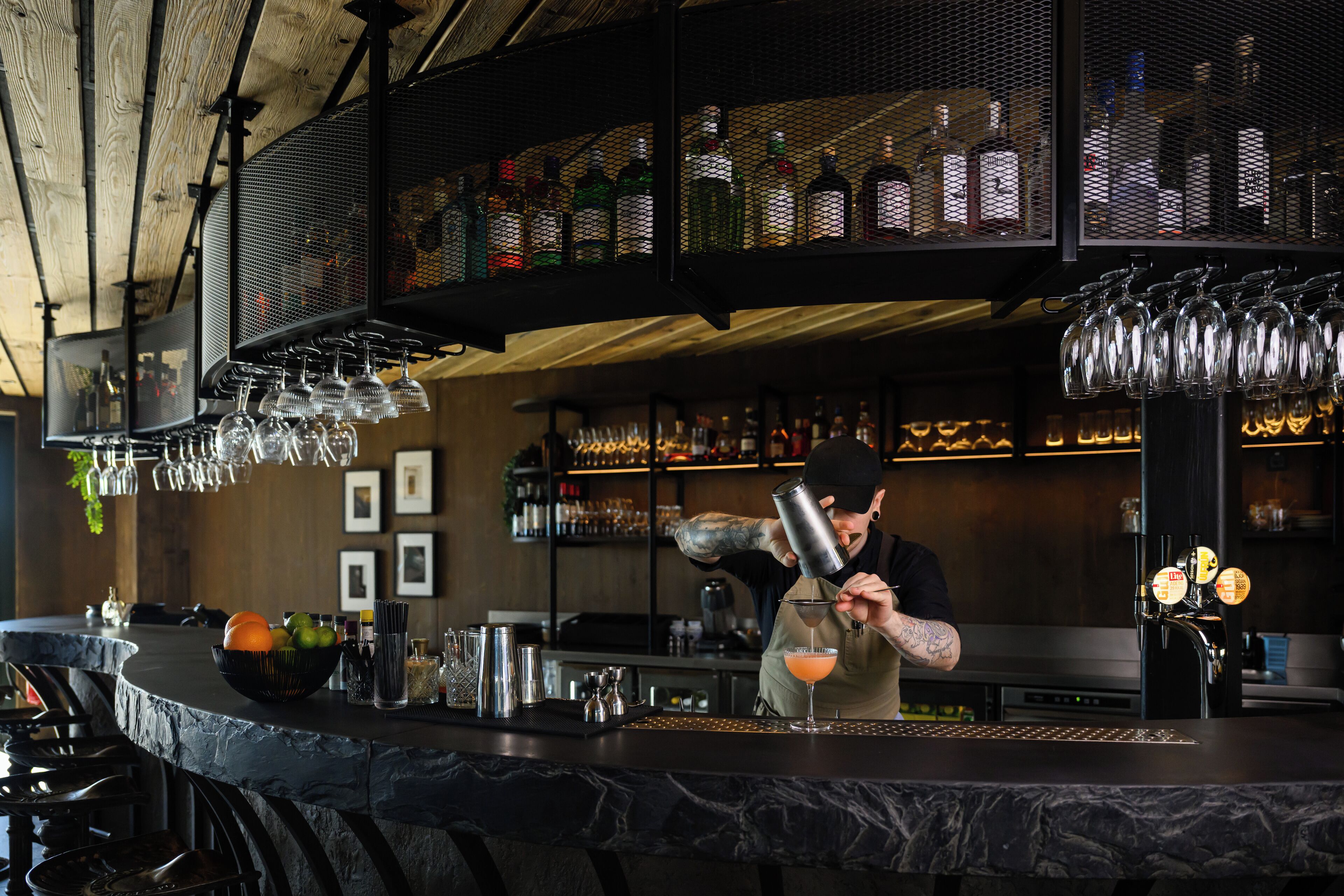 Un espace bar avec un barman préparant un cocktail, des étagères remplies de bouteilles d’alcool et des verres suspendus.