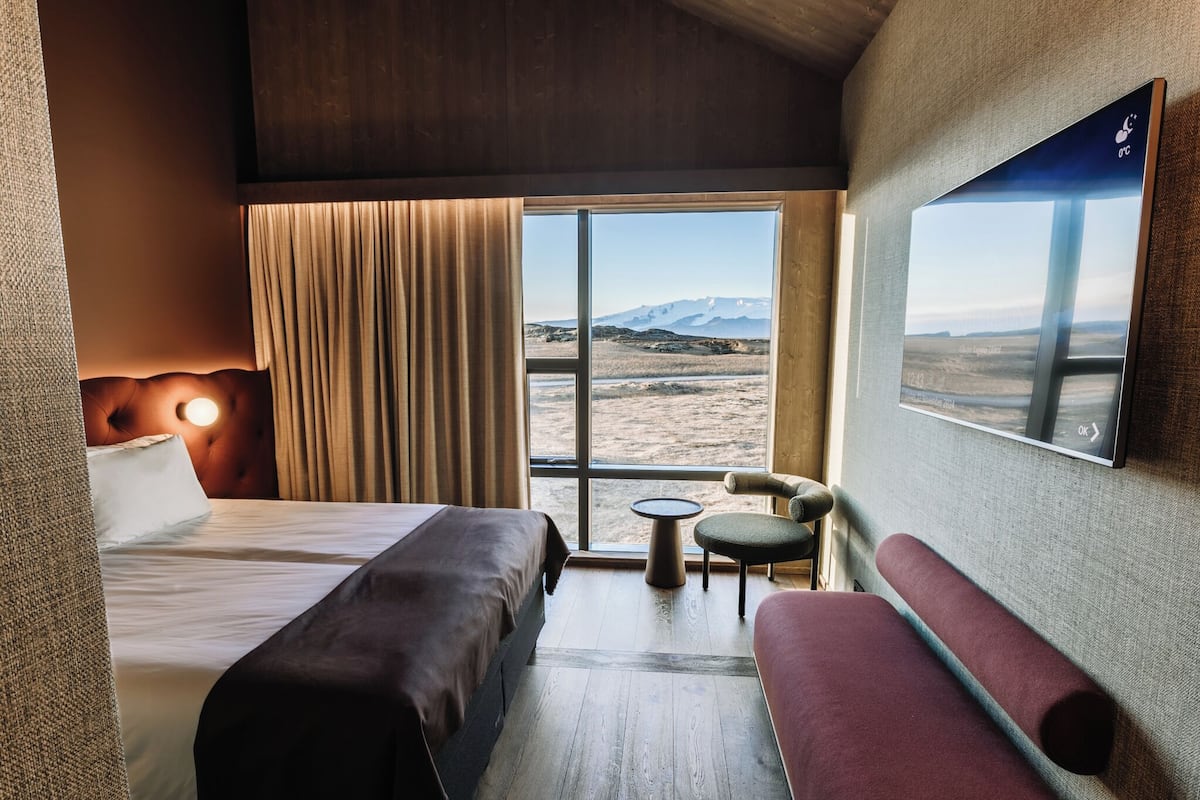 A hotel room with a large window, a bed, a sofa, a small table, and a mounted TV.