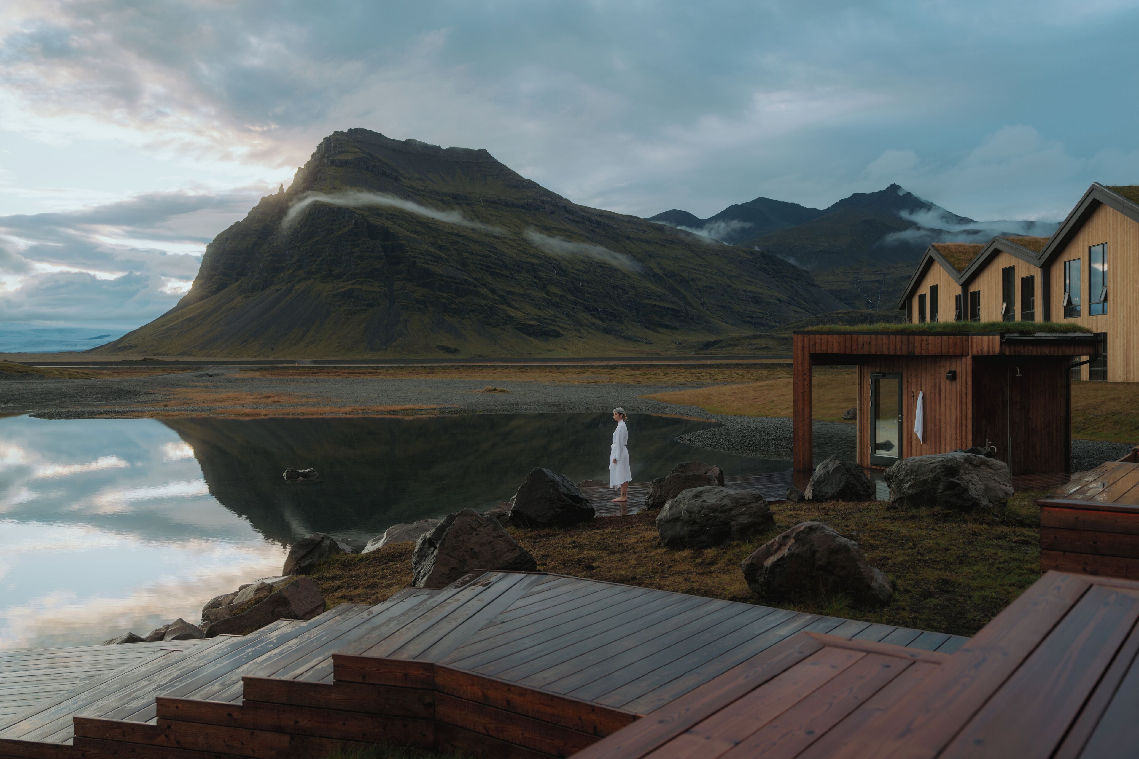 A wooden cabin with a glass door, situated by a calm lake with mountains in the background.