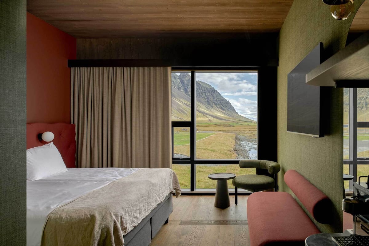 A bedroom with a bed, a small table, a chair, and a sofa. There is a large window with a view of a mountainous landscape.