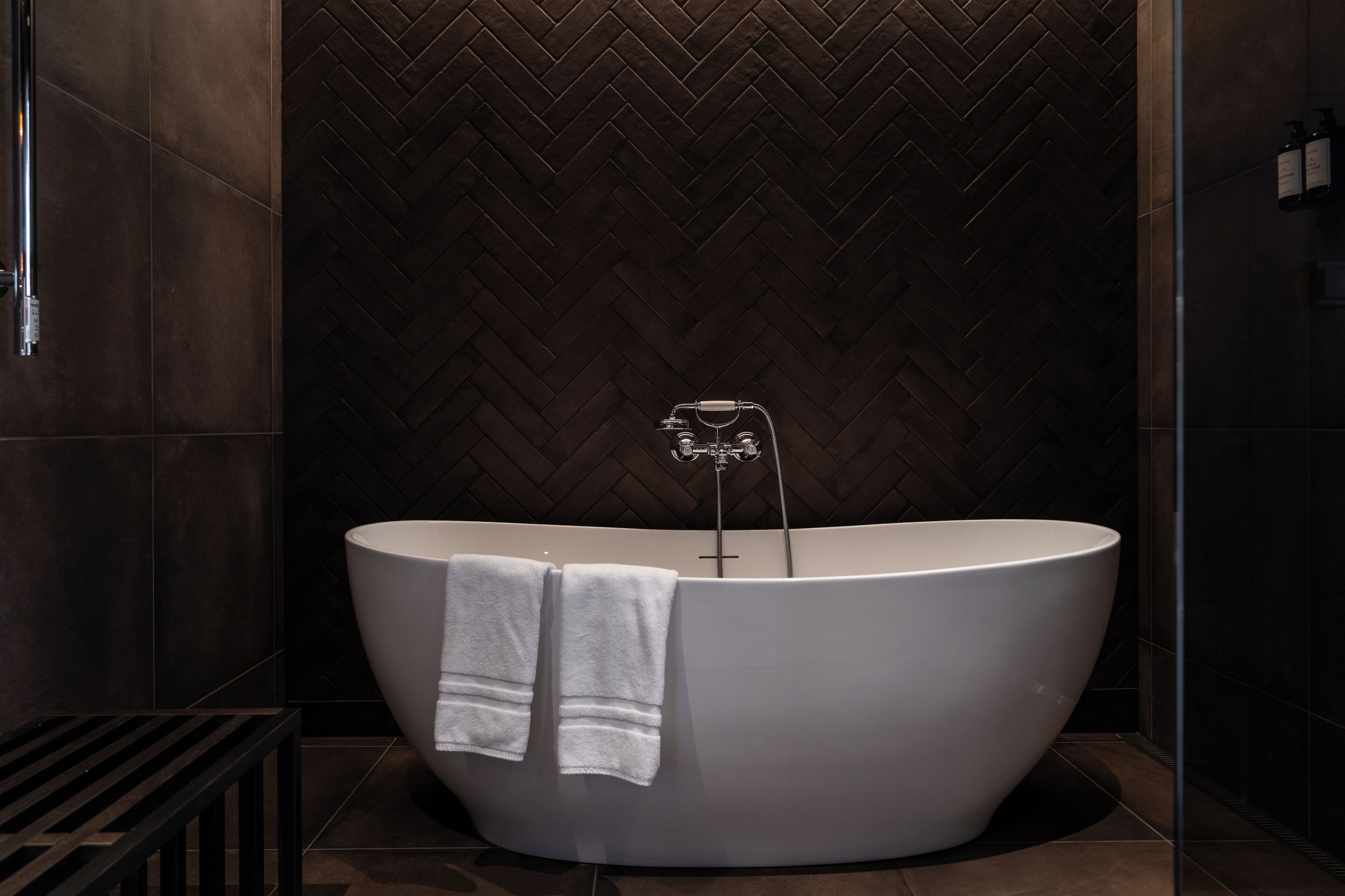 Een moderne badkamer met een vrijstaand bad, een wand met een herringbone-patroon en een metalen kraan.