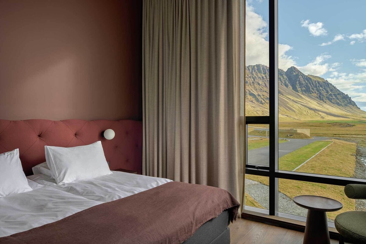 A hotel room with a large bed, a view of mountains through a window, and a small table by the window.