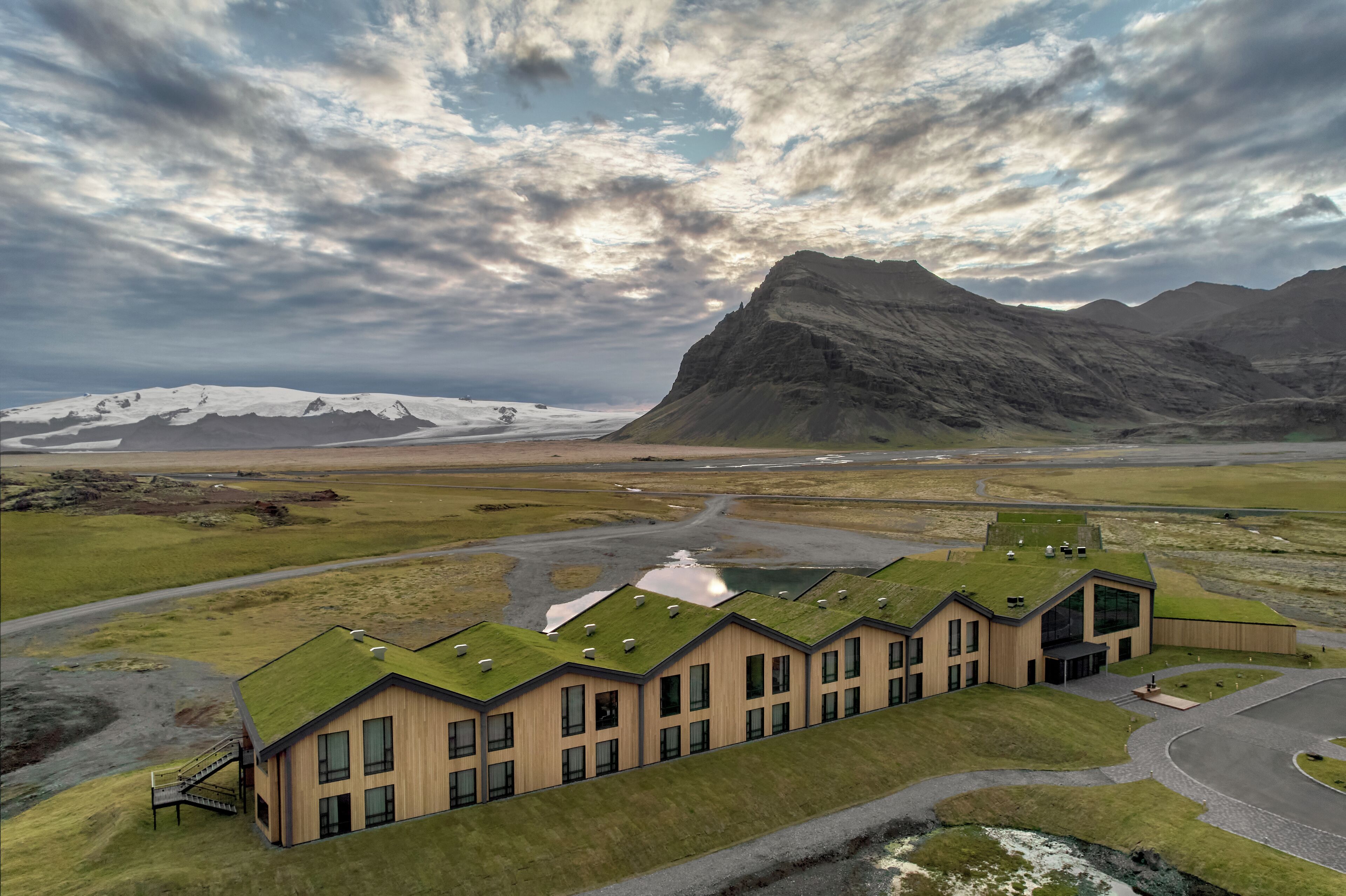 A modern building with a green roof, situated in a rural landscape with mountains in the background.