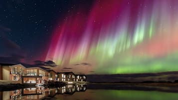 A modern hotel with a reflection in a calm body of water, under a night sky with the Northern Lights.
