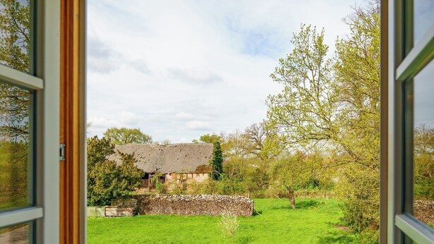 A room with a large window offering a view of a green field and a building with a thatched roof. - Alandblick Inn (Wanzer)