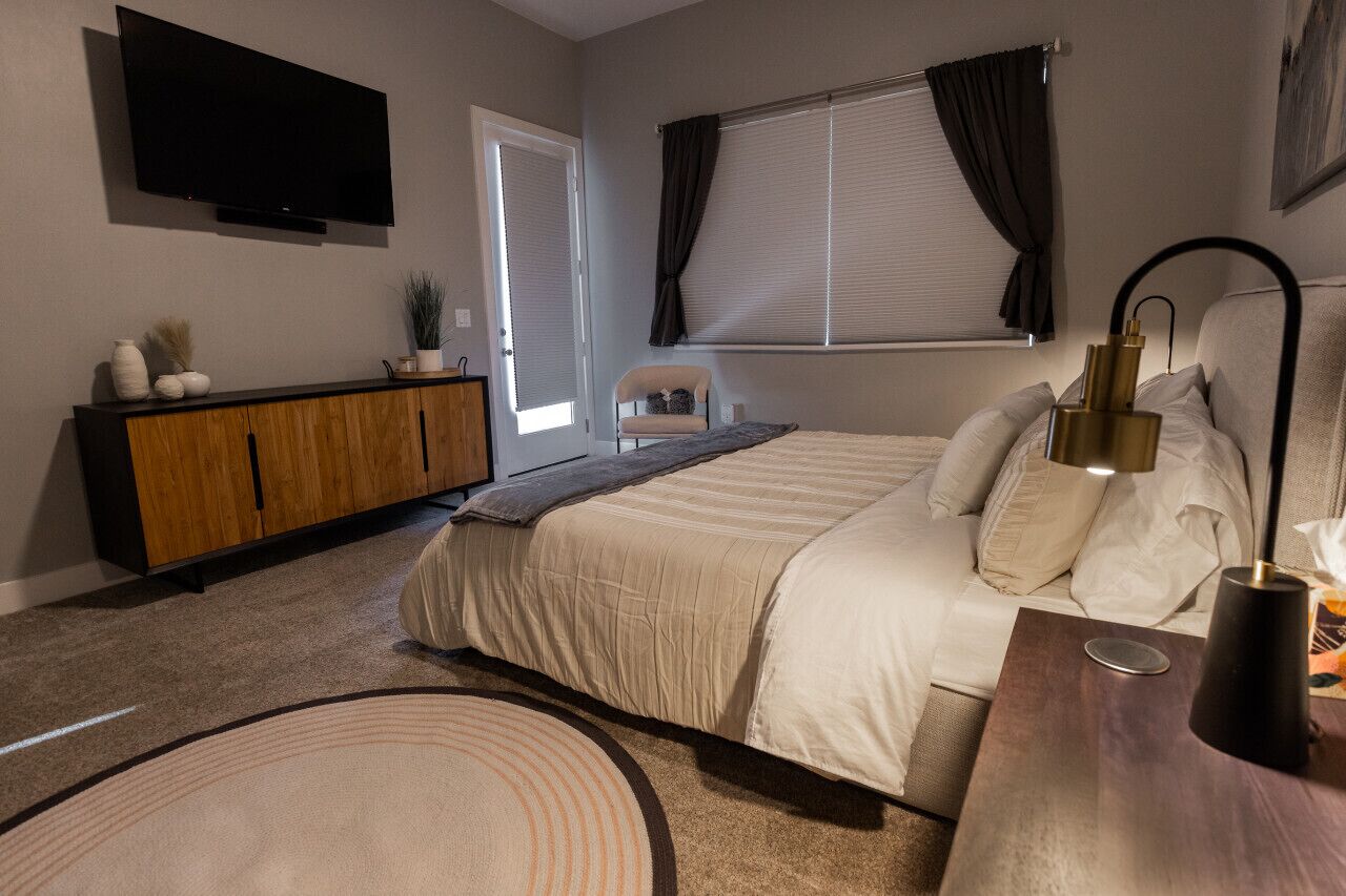 Une chambre à coucher avec un lit, une télévision, une armoire en bois, une chaise et une fenêtre avec des rideaux.