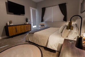 Un dormitorio con cama, televisor montado en la pared, ventana con cortinas, mesita de noche con lámpara y un armario de madera.