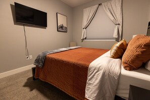 Un dormitorio con cama, televisor montado en la pared, ventana con cortinas e imagen decorativa en la pared.