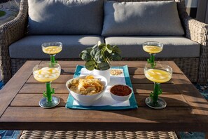 A table set with three drinks, a bowl of chips, and a bowl of salsa, placed on a wooden table outdoors with a wicker sofa in the background. - Par-Tee Timers Paradise by J and Amy BL105619026 (Mesquite)