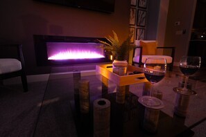 Una sala de estar moderna con chimenea, una mesa de centro de vidrio con copas de vino, una planta de helecho y un sofá.