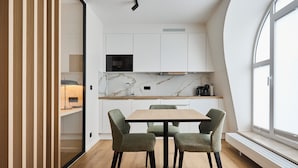 A modern kitchen with white cabinets, a marble backsplash, and a built-in microwave. There is a dining table with four chairs and a bench along the window. - Georges Henri Aparthotel (Brussels)