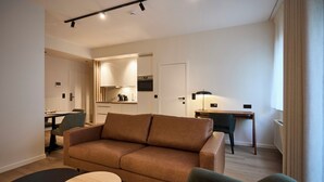 A modern living room with a brown sofa, a round coffee table, and a dining area with a table and chairs. - Georges Henri Aparthotel (Brussels)