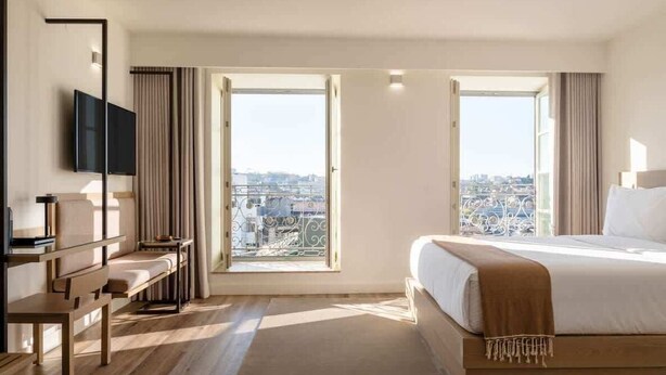 Une chambre d’hôtel avec un grand lit, un bureau, une télévision et une vue sur la ville, visible à travers deux fenêtres.