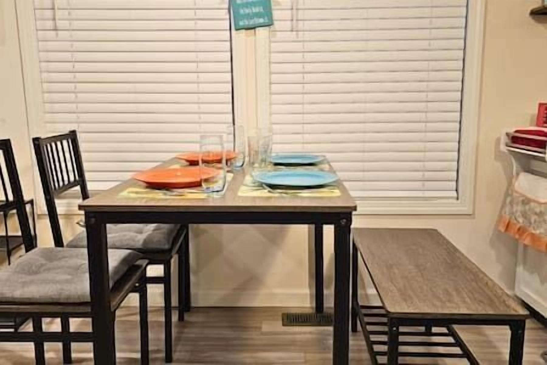 A dining table set with plates and glasses, chairs, and a window with blinds in the background.