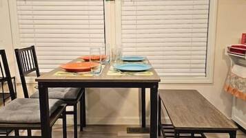 A dining table set with plates and glasses, chairs, and a window with blinds in the background.