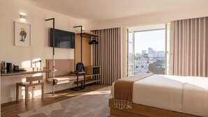 A modern hotel room with a large bed, a desk with a chair, a TV mounted on the wall, and a city view through the window.