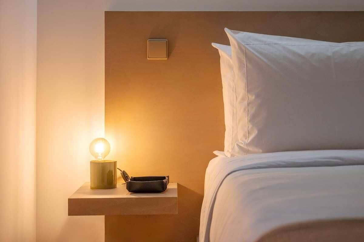 Un lit bien fait, avec du linge de lit blanc et des oreillers, une table de chevet avec une lampe et un plateau noir, et un panneau de commande fixé au mur.