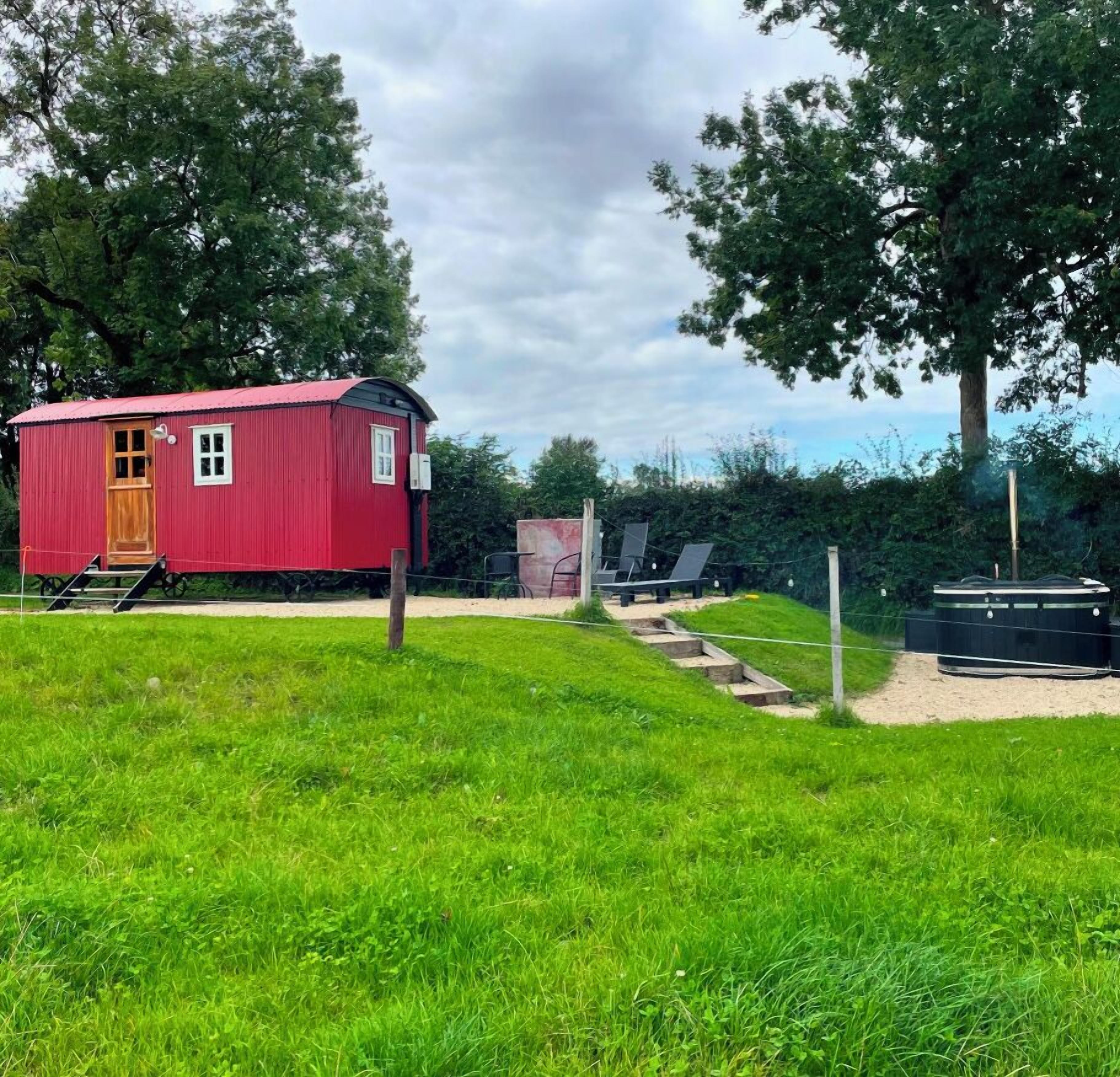 A red cabin with a wooden door and windows, surrounded by a grassy area with trees and a hot tub.
