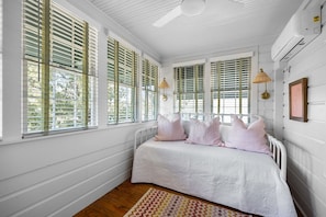 A bedroom with a bed, white walls, wooden flooring, and large windows with shutters.