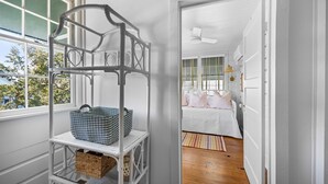 Une pièce avec une étagère métallique blanche contenant des paniers, un lit avec du linge de lit et des oreillers blancs, et une vue sur l’extérieur par une fenêtre.