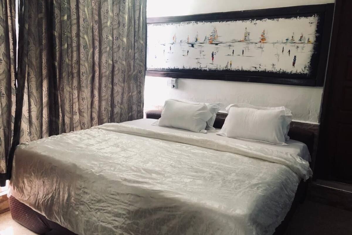A neatly made bed with white linens, a headboard with a painting, and a window with curtains.