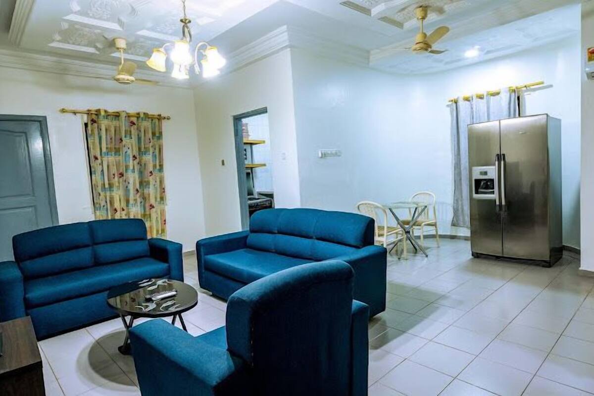 A living room with two blue armchairs, a small round table, a refrigerator, and a dining area with a table and chairs.