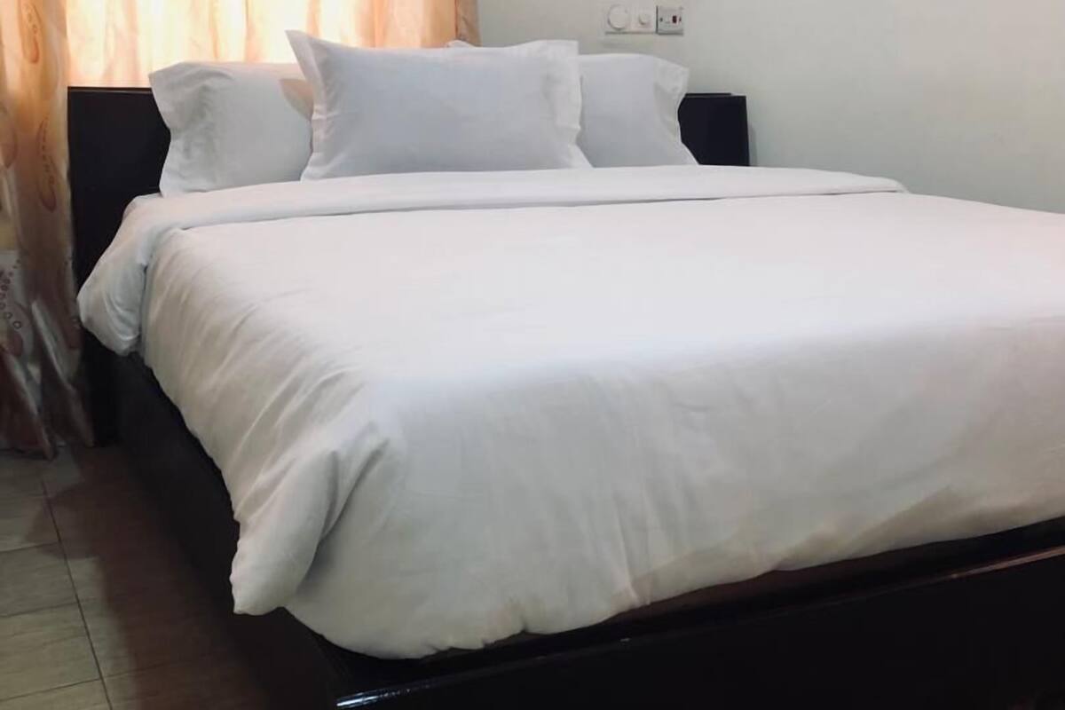 A neatly made bed with white linens in a room with a tiled floor and a curtain.