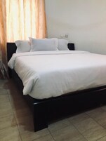 Una cama bien tendida con sábanas blancas en una habitación con suelo de baldosas y una cortina.