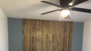 Un dormitorio con una cama, un ventilador de techo, un revestimiento de listones de madera y una mesita de noche con una planta.