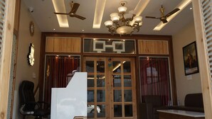 A reception area with a desk, chairs, a chandelier, ceiling fans, and a wooden partition with glass doors. - Maharaja Hotel and Restaurant (Ratia)