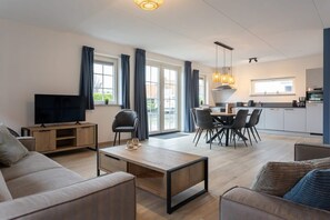 Un salon moderne avec un canapé, une table basse et un téléviseur, donnant sur un coin repas avec des chaises et une cuisine en arrière-plan.