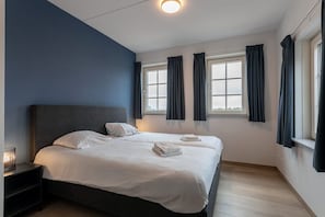 A double bed with white linens, a bedside table with a lamp, and a window with curtains.