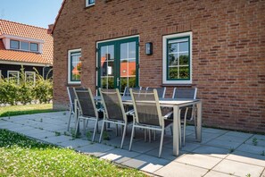 A brick building with a green door and windows, a patio with a table and chairs, and a garden with grass and flowers.