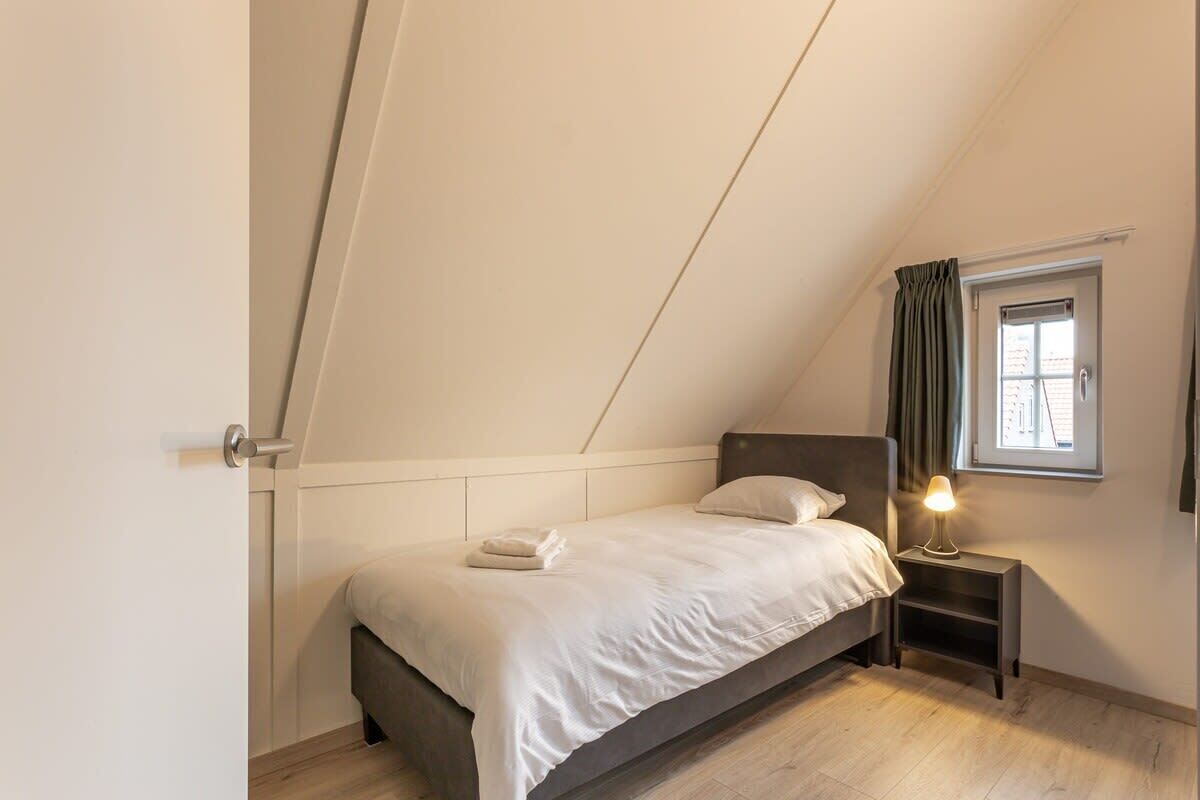 A bedroom with a bed, a bedside table with a lamp, a window with curtains, and a door.