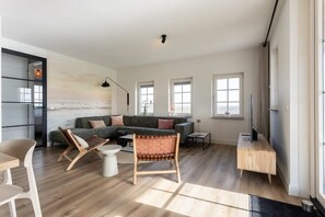 A spacious living room with a sectional sofa, a coffee table, a TV stand, and a large window with a view of the beach. - Holiday home with polder views on Resort Knuitershoek | Knuitershoek 72 (Ossenisse)