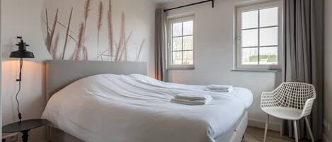 A neatly made bed with white linens, a bedside table with a lamp, a chair, and a wall-mounted artwork.