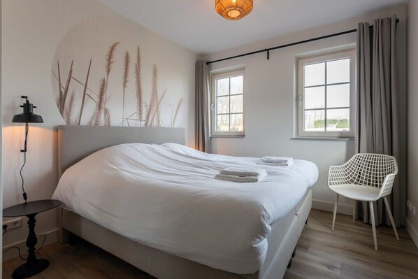 A neatly made bed with white linens, a bedside table with a lamp, a chair, and a wall-mounted artwork. - Holiday home with polder views on Resort Knuitershoek | Knuitershoek 72 (Ossenisse)