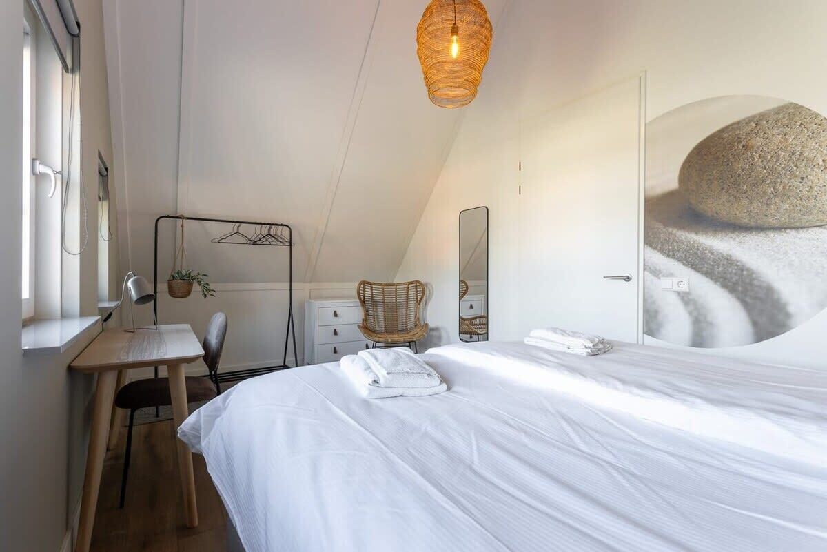 A bedroom with a bed, a desk, a chair, a hanging light fixture, and a large mirror.