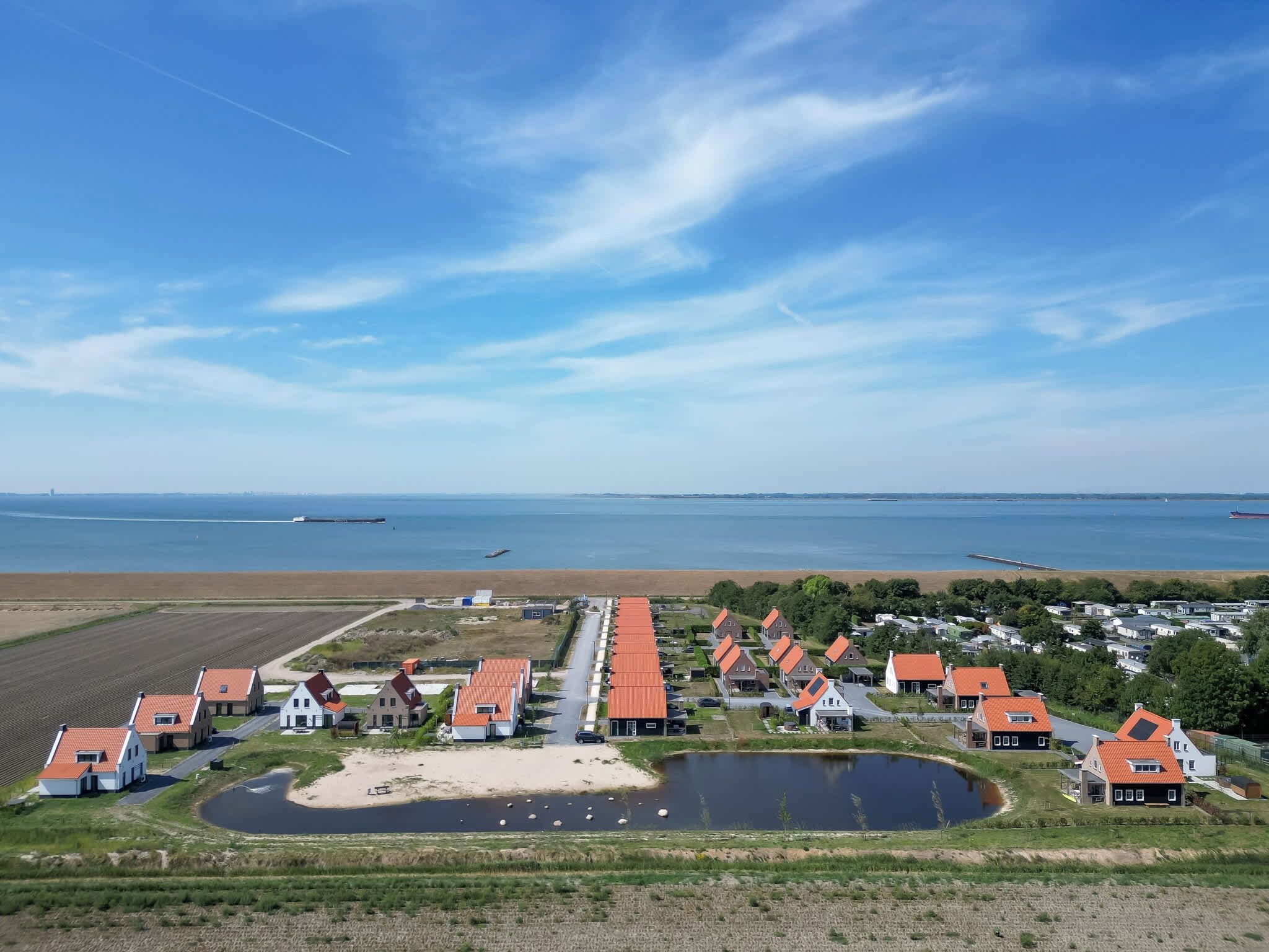 A coastal village with houses featuring red roofs, a small pond, and a view of the sea and distant ships.