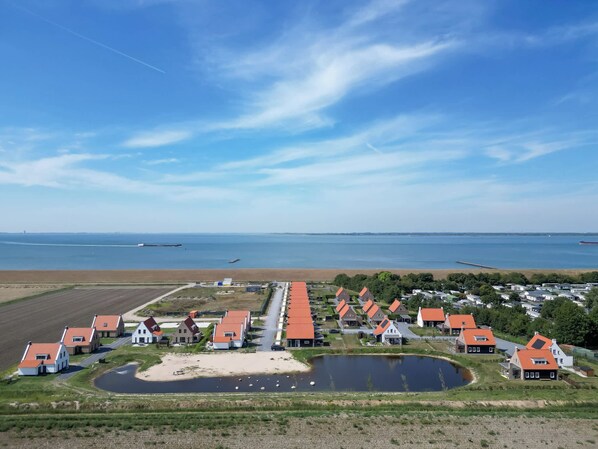A coastal village with houses featuring red roofs, a small pond, and a view of the sea and distant ships.
