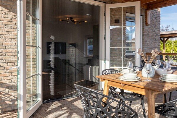 Un área de comedor al aire libre con mesa y sillas de madera, una puerta de cristal que da a un porche cubierto y una pared de ladrillo.