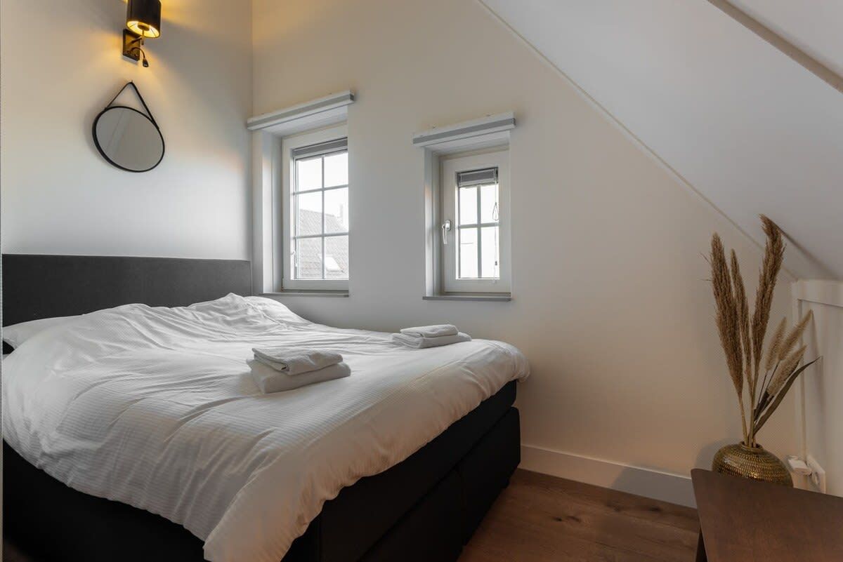 Une chambre à coucher avec un lit, deux fenêtres, un luminaire fixé au mur et un vase contenant des plantes séchées.