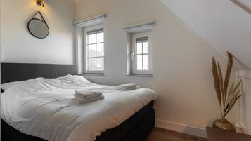 A bedroom with a bed, two windows, a wall-mounted light fixture, and a vase with dried plants.