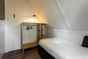 Un dormitorio pequeño y moderno con techo inclinado, una cama con ropa de cama blanca, un armario de madera con estantes y un perchero.